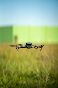 A black drone with four propellers is hovering over a field of tall green grass. In the background, there is a blurred green wall under a clear blue sky.