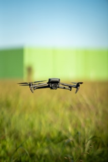 A black drone with four propellers is hovering over a field of tall green grass. In the background, there is a blurred green wall under a clear blue sky.