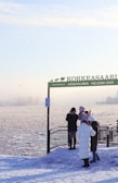A snowy scene with several people dressed in winter clothing, standing near a railing at a snow-covered zoo entrance. The sign reads 'Korkeasaari Högholmen Helsinki Zoo', and there is water in the background with some ice formations. The participants appear to be taking pictures or observing the scenery.