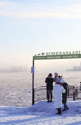 A snowy scene with several people dressed in winter clothing, standing near a railing at a snow-covered zoo entrance. The sign reads 'Korkeasaari Högholmen Helsinki Zoo', and there is water in the background with some ice formations. The participants appear to be taking pictures or observing the scenery.