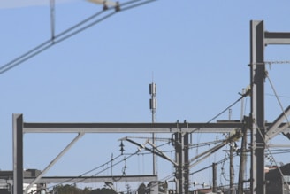 A network of power lines and metal structures is seen against a clear blue sky. In the distance, a telecommunication tower stands tall, surrounded by various wires and poles. The composition creates a geometric pattern.