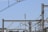 A network of power lines and metal structures is seen against a clear blue sky. In the distance, a telecommunication tower stands tall, surrounded by various wires and poles. The composition creates a geometric pattern.