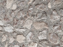 A close-up view of a textured surface consisting of irregularly shaped stones embedded in concrete. The stones vary in shades of light brown and are set against a grayish backdrop, creating a rugged and organic pattern.
