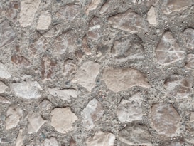 A close-up view of a textured surface consisting of irregularly shaped stones embedded in concrete. The stones vary in shades of light brown and are set against a grayish backdrop, creating a rugged and organic pattern.