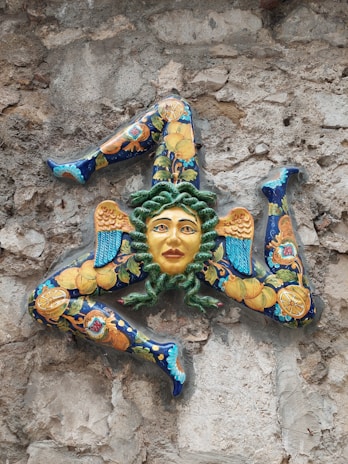 A ceramic artwork featuring a face with green hair surrounded by three bent legs, each embellished with intricate patterns of blue, orange, and yellow. The piece is mounted on a rough stone wall, creating a striking contrast.