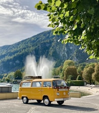 A classic yellow van is parked in a scenic area with a large fountain in the background. The vehicle is adorned with flowers on its front, and a roof rack holds luggage. Surrounding the scene are lush green trees and a mountainous landscape under a partly cloudy sky.