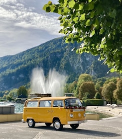A classic yellow van is parked in a scenic area with a large fountain in the background. The vehicle is adorned with flowers on its front, and a roof rack holds luggage. Surrounding the scene are lush green trees and a mountainous landscape under a partly cloudy sky.