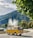 A classic yellow van is parked in a scenic area with a large fountain in the background. The vehicle is adorned with flowers on its front, and a roof rack holds luggage. Surrounding the scene are lush green trees and a mountainous landscape under a partly cloudy sky.