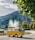 A classic yellow van is parked in a scenic area with a large fountain in the background. The vehicle is adorned with flowers on its front, and a roof rack holds luggage. Surrounding the scene are lush green trees and a mountainous landscape under a partly cloudy sky.