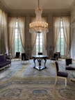A luxurious room features a large, ornate chandelier hanging from the ceiling. Tall, light-colored drapes cover large windows, allowing soft light to fill the space. Elegant chairs with dark upholstery are arranged around a central round table. The room has a richly patterned carpet in muted tones.