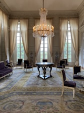 A luxurious room features a large, ornate chandelier hanging from the ceiling. Tall, light-colored drapes cover large windows, allowing soft light to fill the space. Elegant chairs with dark upholstery are arranged around a central round table. The room has a richly patterned carpet in muted tones.