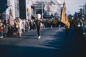 A bustling street scene during a parade features a marching band in uniform, complete with instruments such as trumpets and drums, and majorettes twirling batons. Spectators line both sides of the street, including children and adults, some holding an American flag. Buildings with classic storefront signs, such as 'Mayors Diamond' and 'OSCO Drug', create the backdrop, hinting at a mid-20th-century setting.