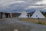 The image features two large white teepees located on a gravel area, with a wooden building nearby. The surroundings include green fields and distant, cloud-covered mountains, creating a rustic and natural setting.