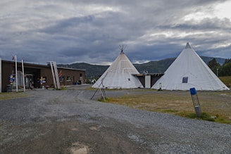 The image features two large white teepees located on a gravel area, with a wooden building nearby. The surroundings include green fields and distant, cloud-covered mountains, creating a rustic and natural setting.