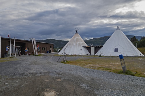 The image features two large white teepees located on a gravel area, with a wooden building nearby. The surroundings include green fields and distant, cloud-covered mountains, creating a rustic and natural setting.