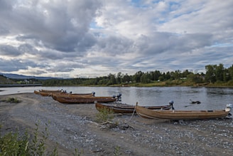 Several wooden boats with motors are lined up on a rocky riverbank, with lush green trees lining the opposite bank. The sky is overcast with thick clouds, suggesting an impending change in weather. The water reflects the gray tones of the sky, and there is a bridge visible in the distance.