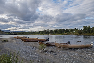 Several wooden boats with motors are lined up on a rocky riverbank, with lush green trees lining the opposite bank. The sky is overcast with thick clouds, suggesting an impending change in weather. The water reflects the gray tones of the sky, and there is a bridge visible in the distance.