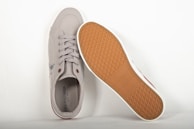 A pair of casual sneakers displayed on a light background. One shoe is positioned upright, showing the laces and the upper part, while the other shoe is flipped to reveal the textured sole. The sneakers are gray with a brown rubber sole.