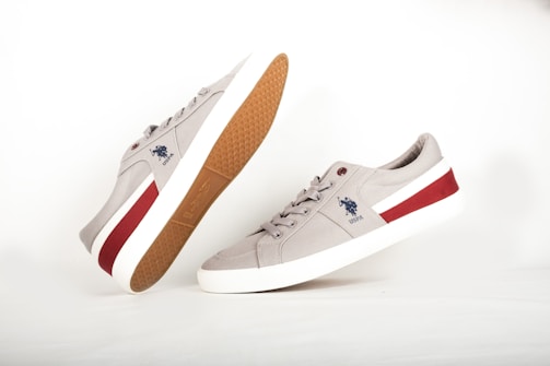 A pair of light gray sneakers displayed against a white background. One shoe is standing upright while the other is tilted, showcasing its light brown sole with a textured pattern. The sneakers feature a white sole with a red accent stripe on the side and a logo of a rider on a horse in blue on the side.