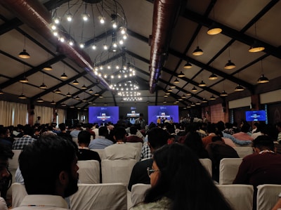 A large group of people seated in a conference hall with high ceilings and industrial-style lighting. The attendees are facing a stage where a presentation is displayed on multiple screens, indicating a technology or community event.
