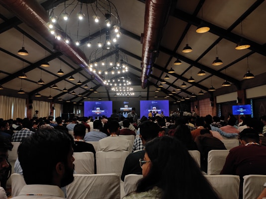 A large group of people seated in a conference hall with high ceilings and industrial-style lighting. The attendees are facing a stage where a presentation is displayed on multiple screens, indicating a technology or community event.