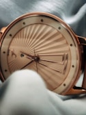 A close-up view of an elegant wristwatch with a gold-tone case and detailing. The watch features a radiant pattern on the dial, gold hour markers, and uniquely designed hour, minute, and second hands. The watch is partially obscured by a soft fabric, adding a sense of luxury and sophistication.