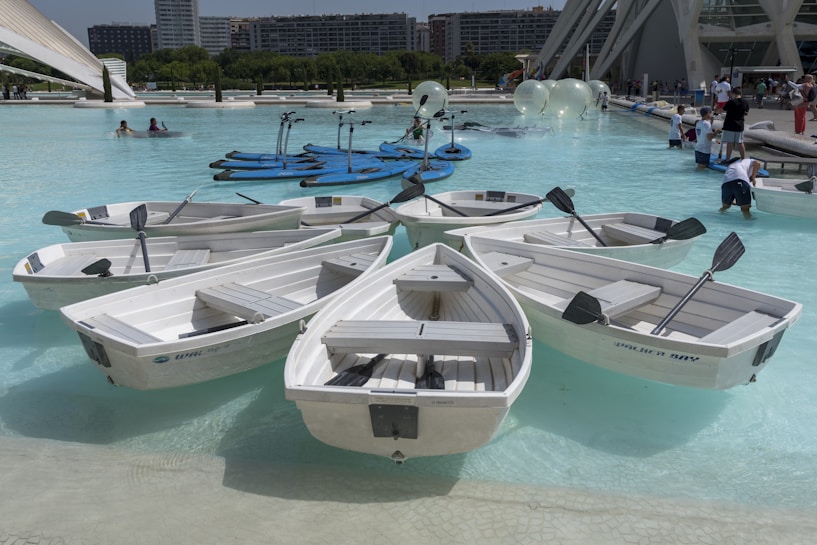 Several white rowboats are grouped together in a pool of clear blue water. Nearby, people can be seen engaging in water activities, and in the background, there are tall buildings and a modern architectural structure. The scene is lively and appears to be a part of an urban recreational area.