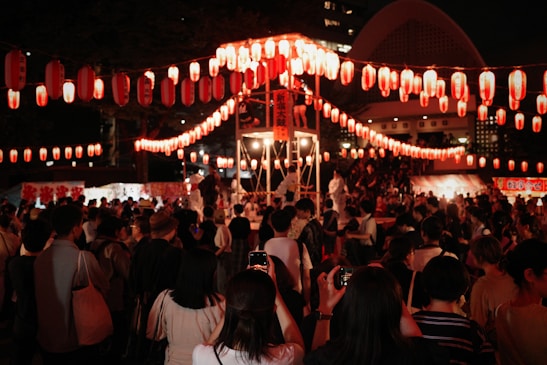 A lively festival scene featuring a large crowd of people gathered at night. The area is illuminated with numerous red lanterns hung in strings overhead, creating a festive and warm atmosphere. A stage is visible in the background with performers dressed in white, and various stalls or booths line the periphery.