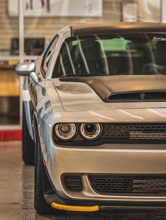 A sleek modern car viewed from the front, captured indoors with a reflective surface highlighting its polished body. The vehicle features dual round headlights, a ventilated hood, and a prominent grille, conveying a powerful and robust appearance. Background elements are slightly blurred, focusing attention on the car.