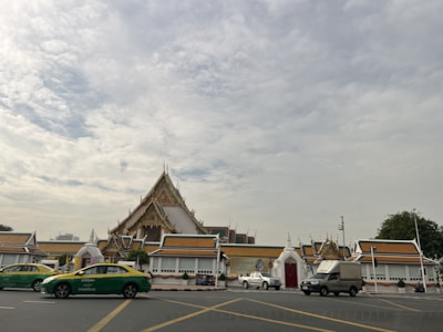 A traditional temple with ornate architecture and pointed roofs set against a cloudy sky. Several vehicles, including green and yellow taxis, are seen on the road in front of the temple. Trees and a modern building are visible in the distance.