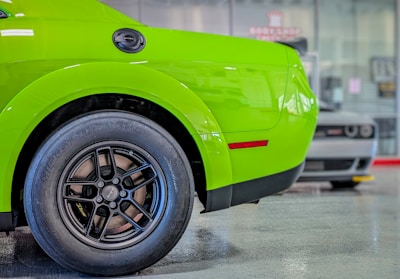 A bright green car is prominently displayed with focus on the rear section and tire. The background shows another car slightly out of focus in a showroom setting, with the floor reflecting some light.