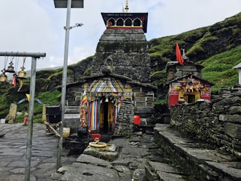 An ancient stone temple adorned with colorful flags and intricate decorations. The temple is situated in a mountainous area with lush greenery surrounding it. Bells hang from poles in the foreground, adding a traditional and spiritual ambiance to the scene. The architecture is rustic, with stone steps leading to the entrance, and a small red shrine is visible to the side.