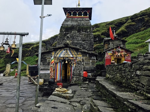 An ancient stone temple adorned with colorful flags and intricate decorations. The temple is situated in a mountainous area with lush greenery surrounding it. Bells hang from poles in the foreground, adding a traditional and spiritual ambiance to the scene. The architecture is rustic, with stone steps leading to the entrance, and a small red shrine is visible to the side.