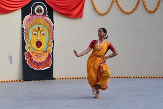 A woman dressed in traditional Indian attire is performing a classical dance in front of a painted backdrop depicting a colorful face with ornate features. The scene is set against a decorated wall adorned with a red fabric and floral garlands.