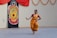 A woman dressed in traditional Indian attire is performing a classical dance in front of a painted backdrop depicting a colorful face with ornate features. The scene is set against a decorated wall adorned with a red fabric and floral garlands.