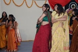 A group of women are dressed in colorful traditional Indian sarees, engaging in a celebratory dance. The background is adorned with hanging marigold garlands and a decorative red drape. The women appear joyful and are smiling as they perform.
