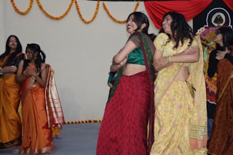 A group of women are dressed in colorful traditional Indian sarees, engaging in a celebratory dance. The background is adorned with hanging marigold garlands and a decorative red drape. The women appear joyful and are smiling as they perform.