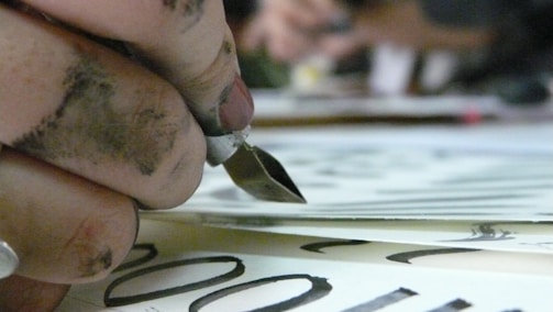 A close-up of a hand holding a calligraphy pen, with fingers stained by ink. The pen is poised above paper with elegant black lettering visible. The focus is on the pen and the inky hand, while the background is blurred.