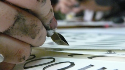 A close-up of a hand holding a calligraphy pen, with fingers stained by ink. The pen is poised above paper with elegant black lettering visible. The focus is on the pen and the inky hand, while the background is blurred.