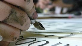 A close-up of a hand holding a calligraphy pen, with fingers stained by ink. The pen is poised above paper with elegant black lettering visible. The focus is on the pen and the inky hand, while the background is blurred.