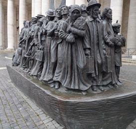 A bronze sculpture depicting a group of people standing closely together, each adorned in varied historical attire suggesting different eras and cultures, symbolizing migration or a journey. Their expressions are solemn and determined, with some holding suitcases and others carrying children.