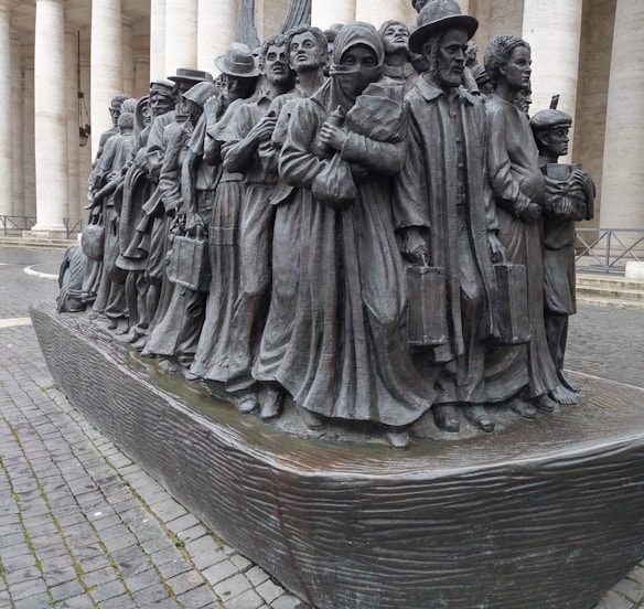 A bronze sculpture depicting a group of people standing closely together, each adorned in varied historical attire suggesting different eras and cultures, symbolizing migration or a journey. Their expressions are solemn and determined, with some holding suitcases and others carrying children.