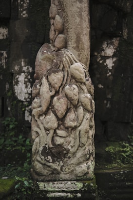A detailed stone carving featuring an intricate design with intertwining shapes, likely representing flora or abstract forms. The carving is weathered, showcasing a combination of smooth and textured surfaces with natural patina. Green foliage and moss are seen growing around the base of the structure, suggesting a setting in nature or an ancient ruin.
