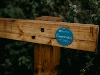 A wooden fence post features a circular green sign with text indicating 'Green Way' and mentions 'spaces for people and wildlife.' It is secured with small nails and depicted against a backdrop of dense green foliage.