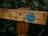 A wooden fence post features a circular green sign with text indicating 'Green Way' and mentions 'spaces for people and wildlife.' It is secured with small nails and depicted against a backdrop of dense green foliage.