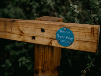 A wooden fence post features a circular green sign with text indicating 'Green Way' and mentions 'spaces for people and wildlife.' It is secured with small nails and depicted against a backdrop of dense green foliage.