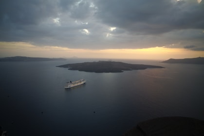 A large cruise ship sails on a vast expanse of calm, dark blue sea during a serene sunset. The sky is filled with dense, grey clouds that obscure some of the light, creating a dramatic atmosphere. In the distance, several dark islands sit silhouetted against the horizon, where the sun casts a warm, golden glow that gradually fades into the cooler tones of the evening.