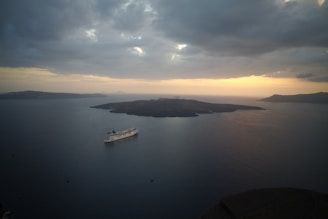 A large cruise ship sails on a vast expanse of calm, dark blue sea during a serene sunset. The sky is filled with dense, grey clouds that obscure some of the light, creating a dramatic atmosphere. In the distance, several dark islands sit silhouetted against the horizon, where the sun casts a warm, golden glow that gradually fades into the cooler tones of the evening.