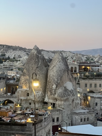 Unique rock formations resembling pointed towers rise prominently against a backdrop of rolling hills. These formations are part of a small town built with stone structures. Several terraces and buildings are visible, with people dining outdoors under subtle lighting, creating a cozy atmosphere.