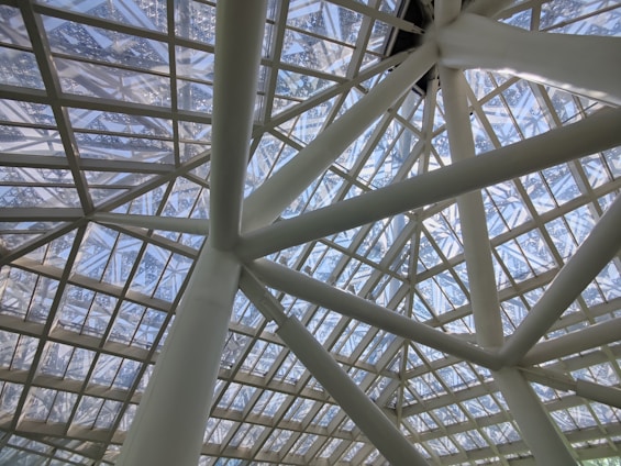 A geometric ceiling structure featuring intersecting metal beams and glass panels. The ceiling showcases a modern architectural design, allowing natural light to filter through the transparent sections.